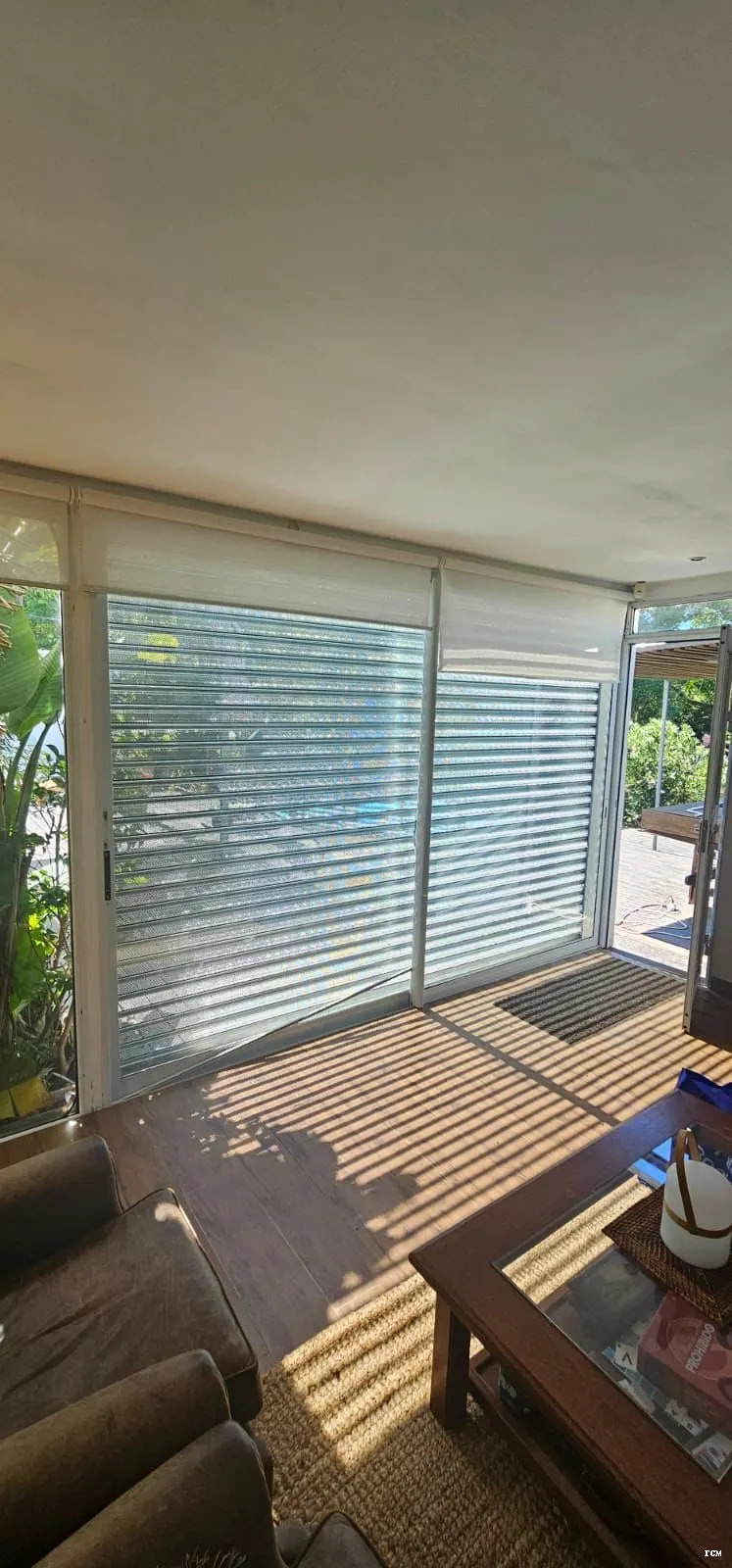 Interior de un living con puertas correderas de persianas metálicas microperforadas, mesa de madera y sofá bajo luz solar.