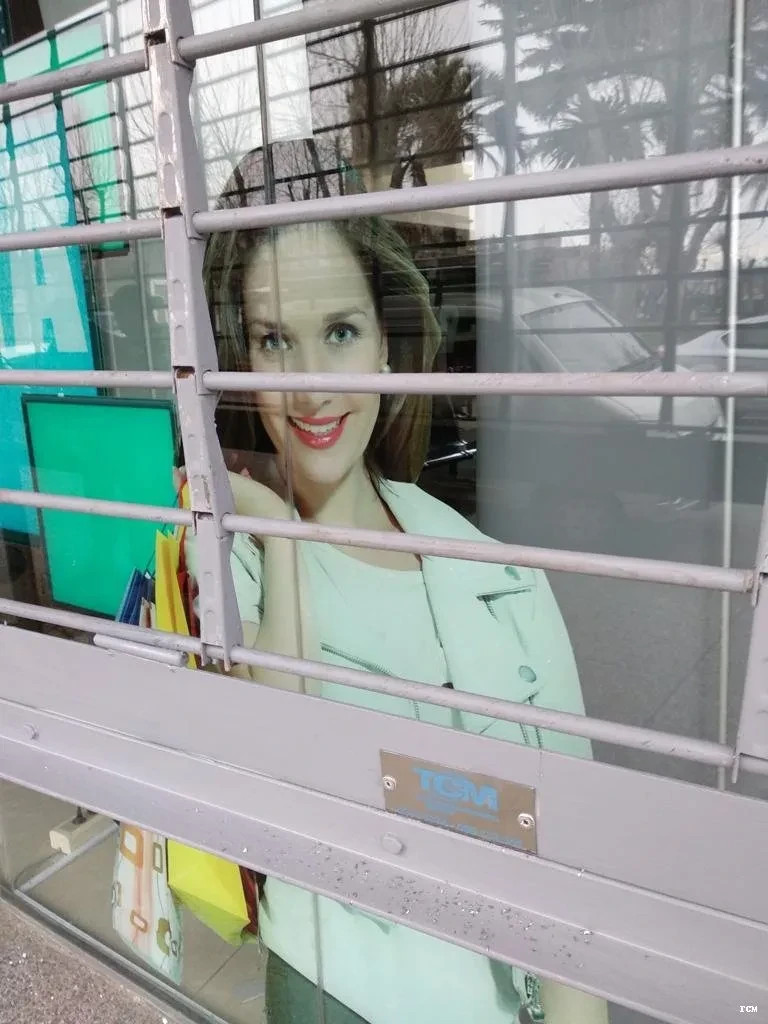 Mujer sonriente asomada detrás de una reja metálica, con oficina moderna y letrero ICM visible.